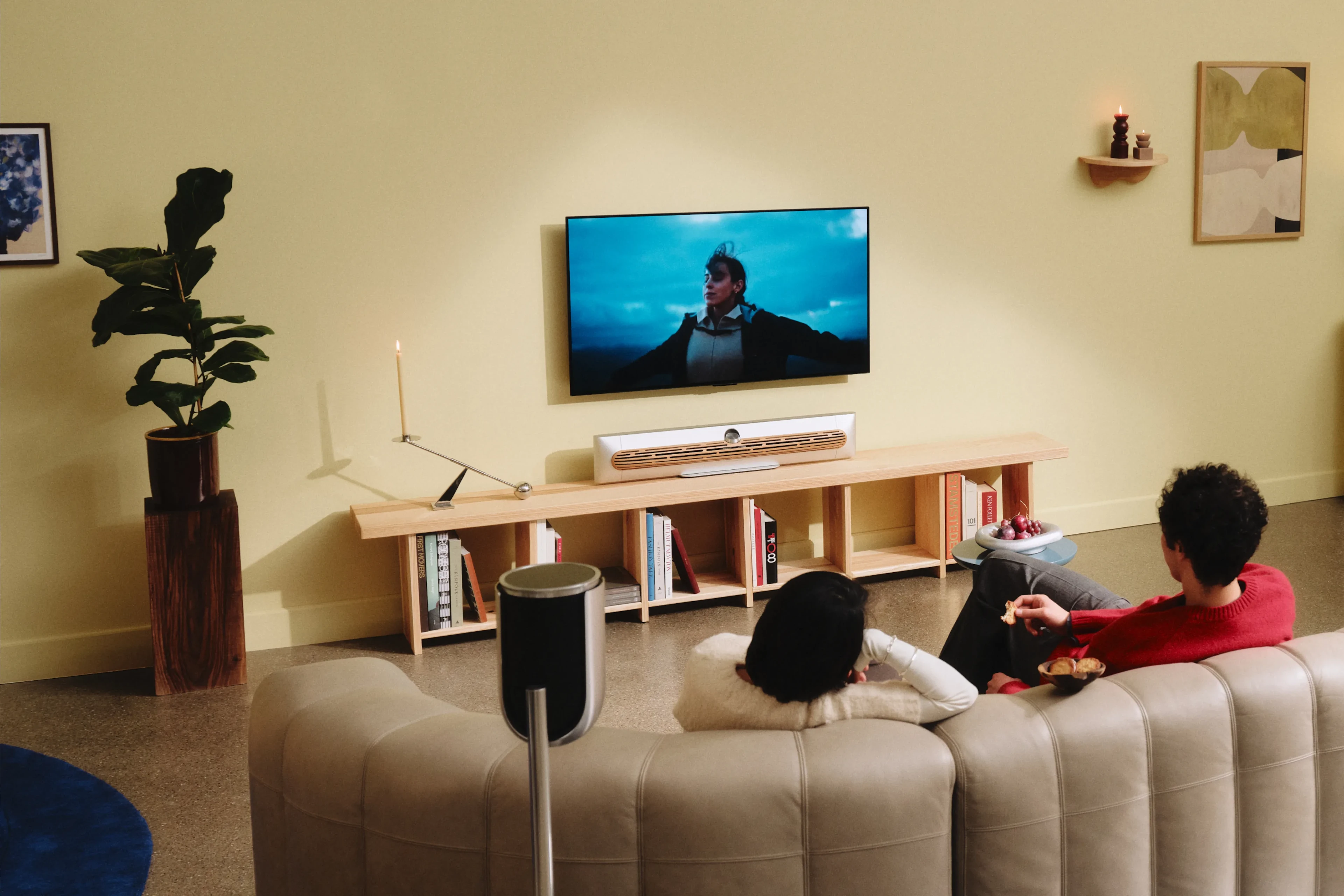 A couple sitting on a sofa in a modern interior living room watching TV, and there is Beosound Premiere Natural aluminium soundbar below the TV.