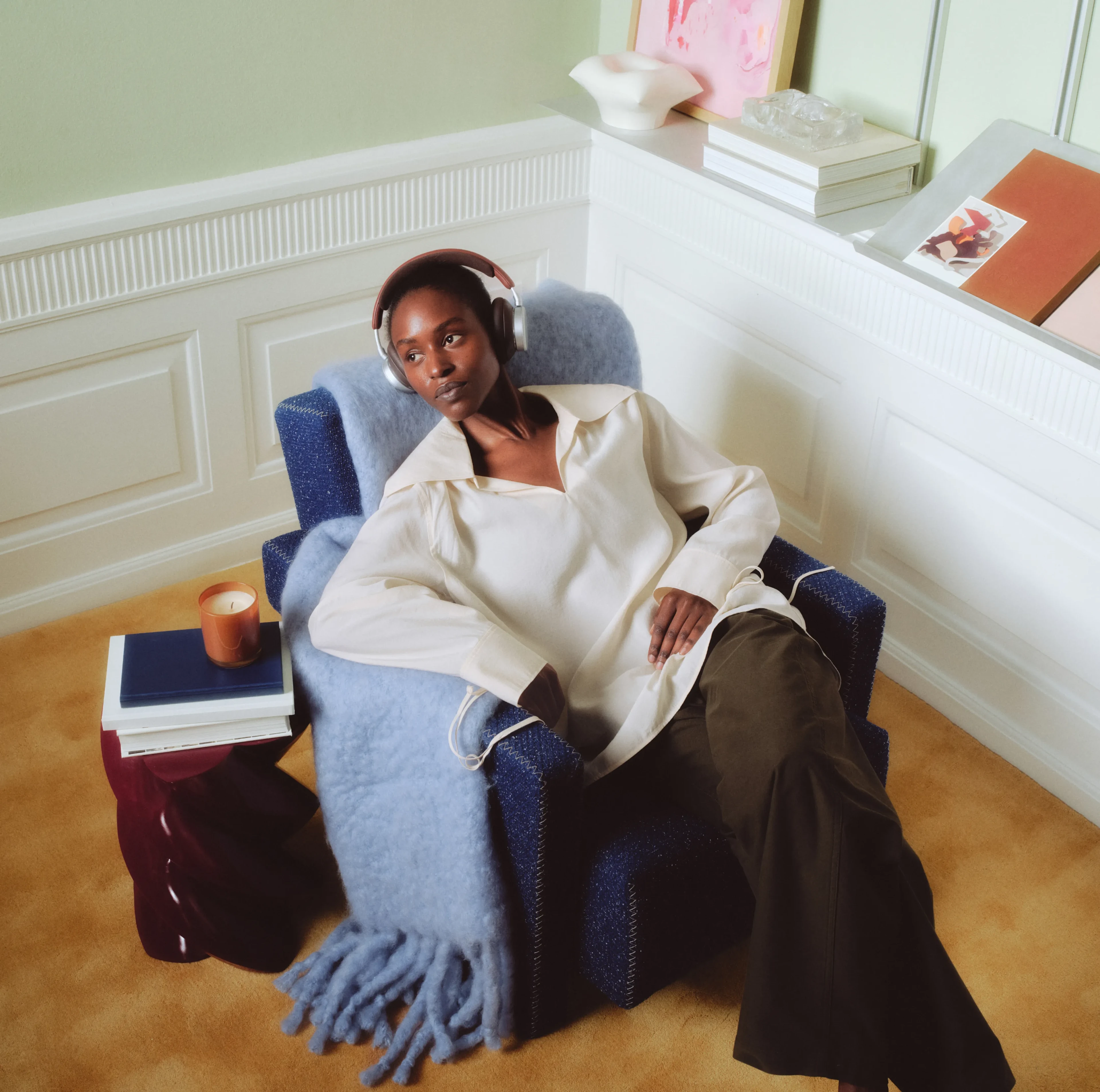Person relaxing in blue armchair wearing white shirt, Beoplay H100 headphones, with books and candle nearby in cozy room setting.