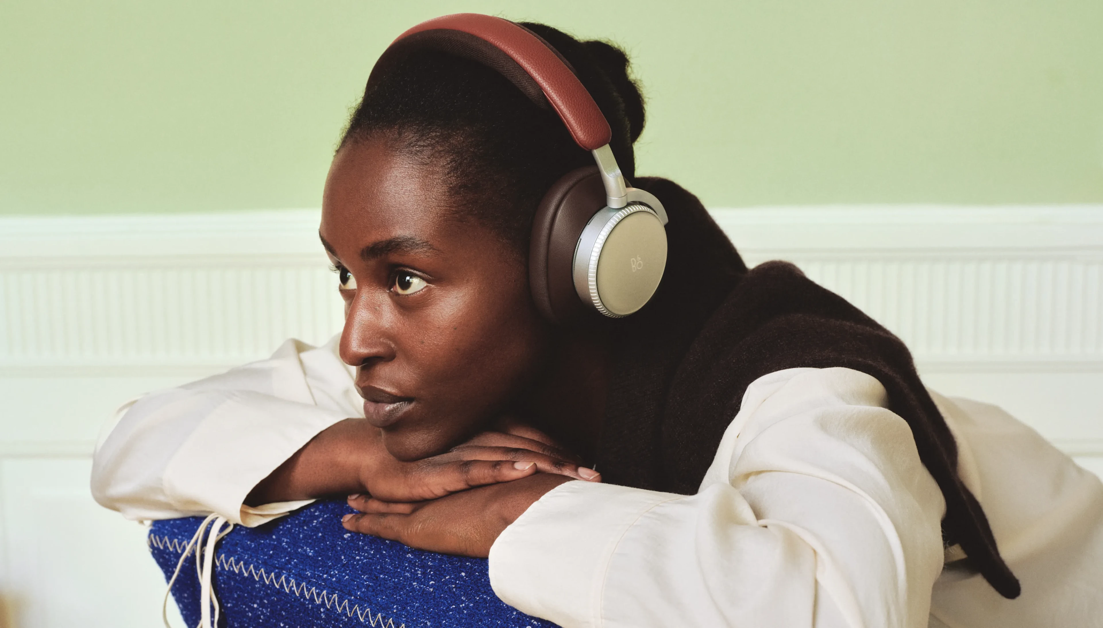 Person wearing stylish over-ear Beoplay H100 Centennial Collection headphones with red headband, resting on blue cushion against light green wall.