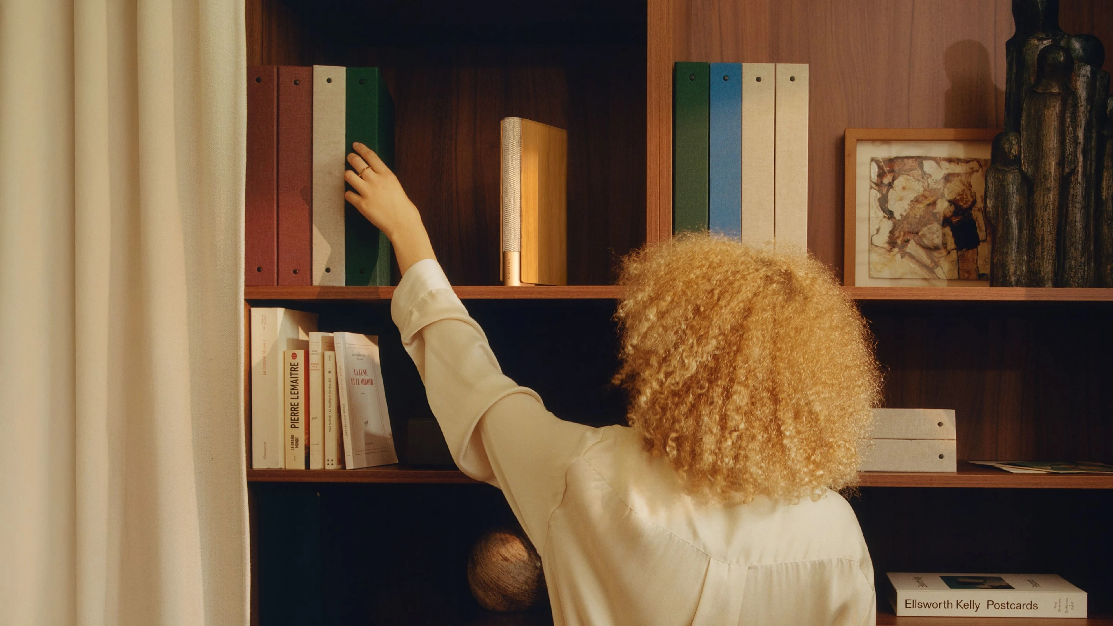 Gold tone Beosound Emerge on a shelf with other books and a lady is trying to pull out one of the books