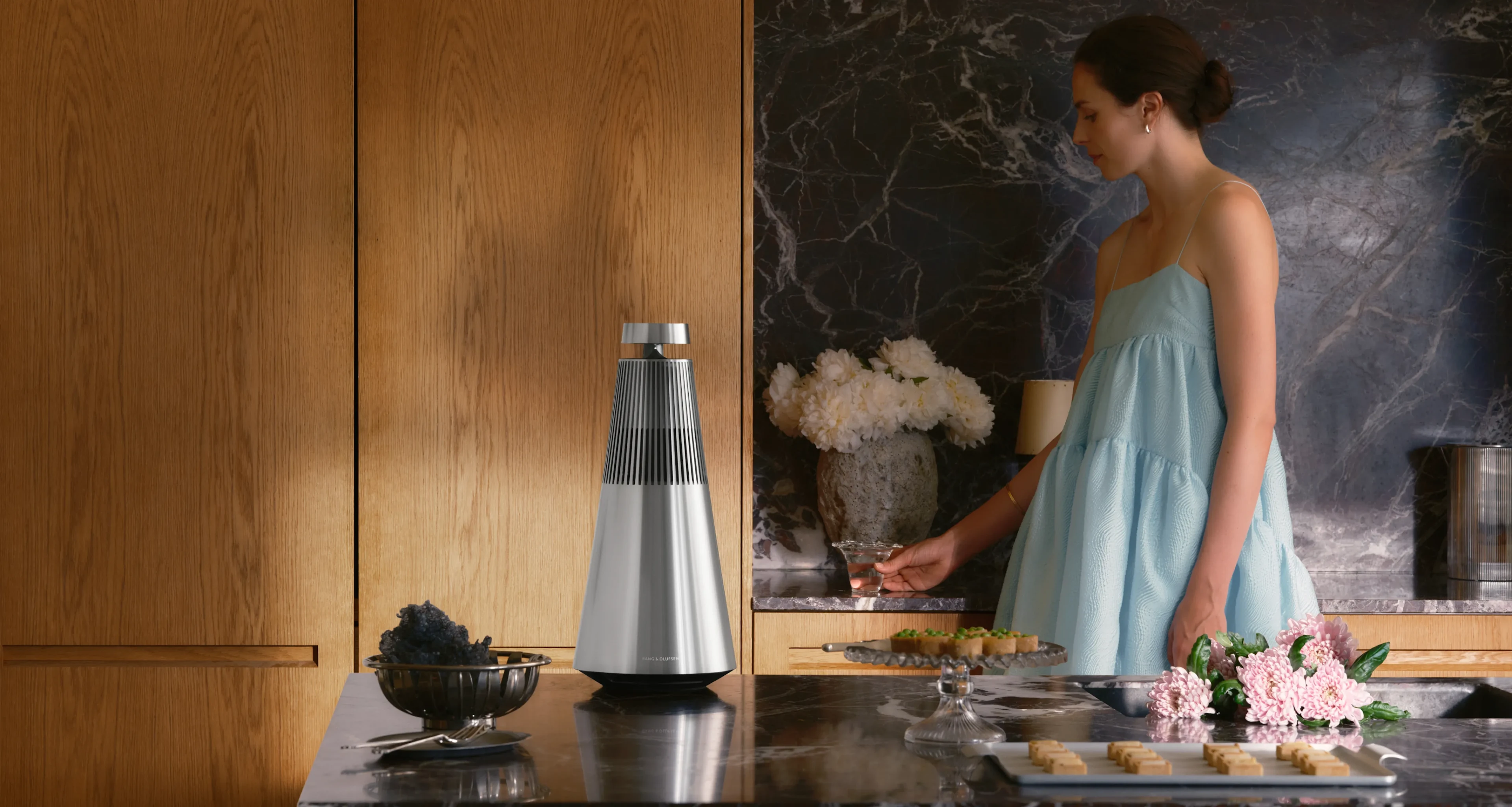 Una mujer con un vestido azul fluido en la cocina, junto a un altavoz Beosound 2 colocado sobre la encimera