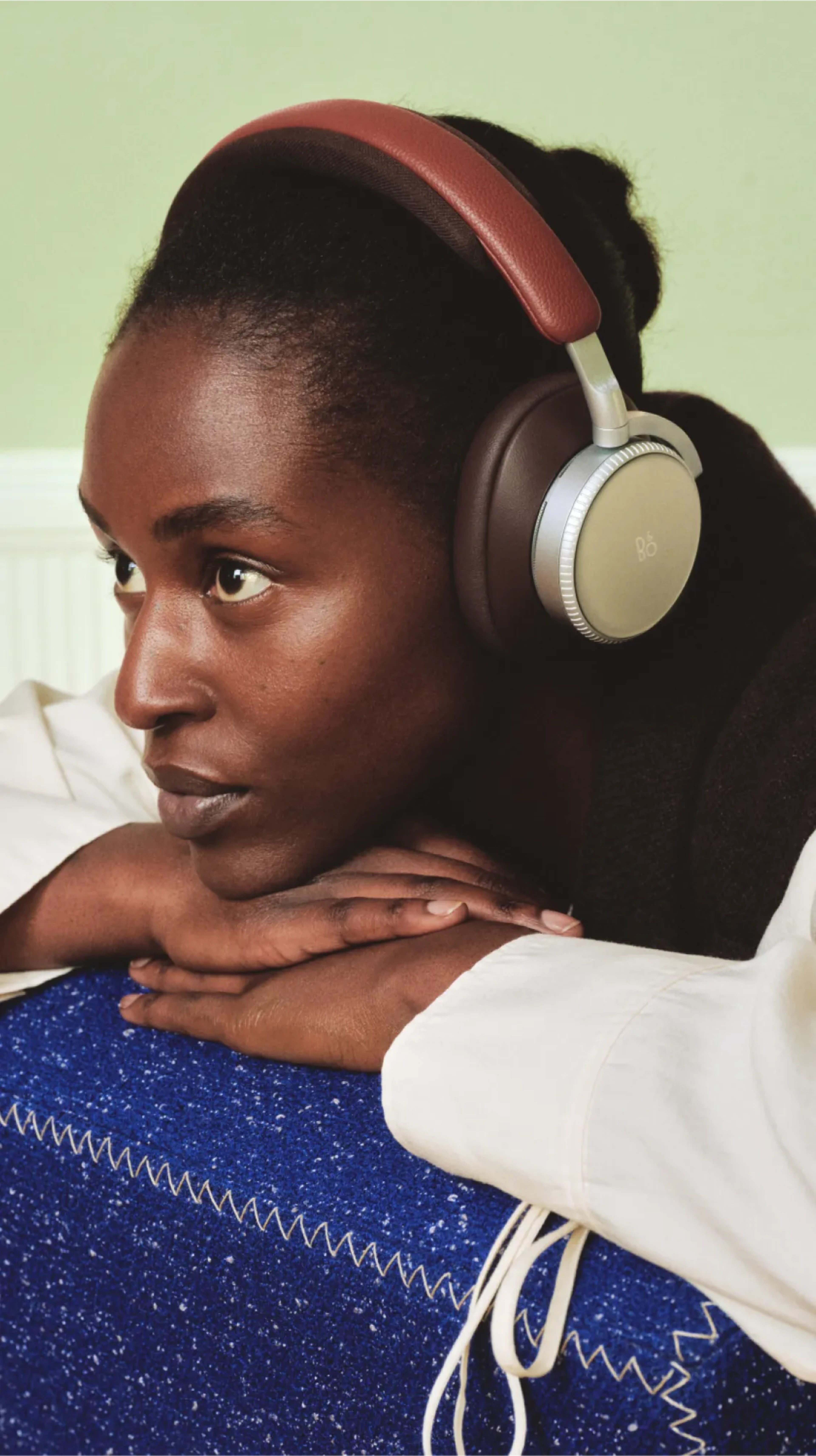 Person wearing stylish over-ear Beoplay H100 Centennial Collection headphones with red headband, resting on blue cushion against light green wall.