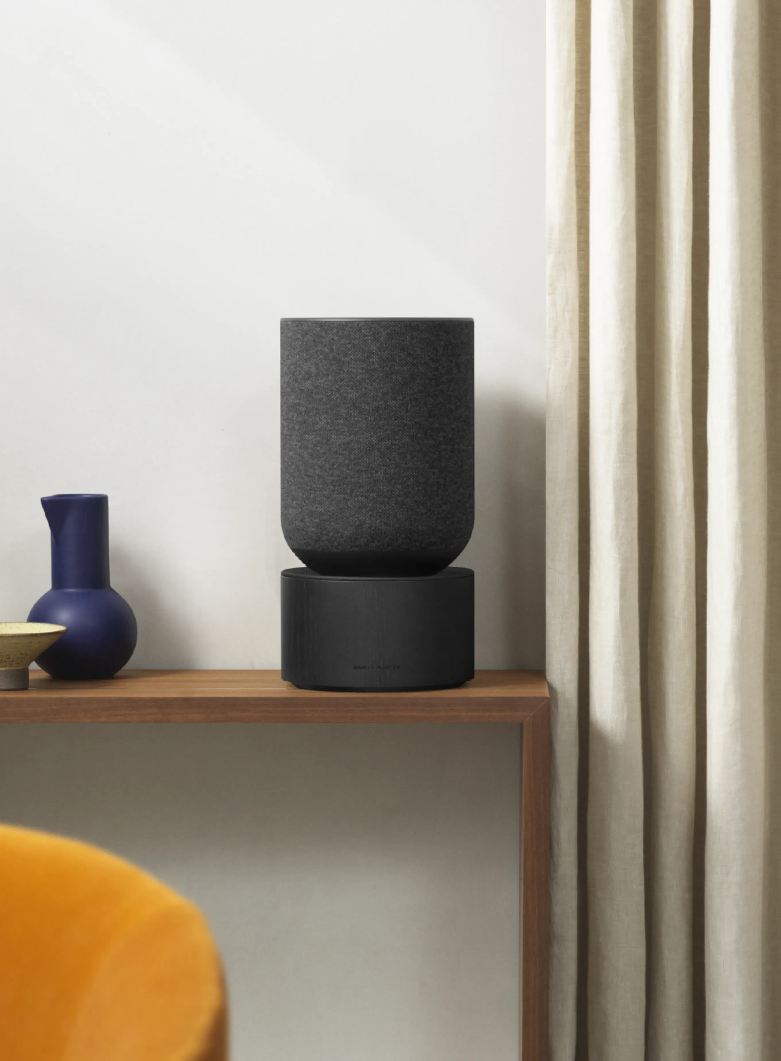Modern living room with wooden console displaying blue vase, black Beosound Balance speaker, and decorative items beside cream curtains.