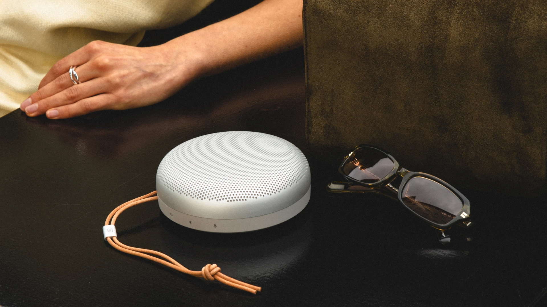 Beosound A1 3rd gen natural aluminium color on a table next to a sunglass and a bag