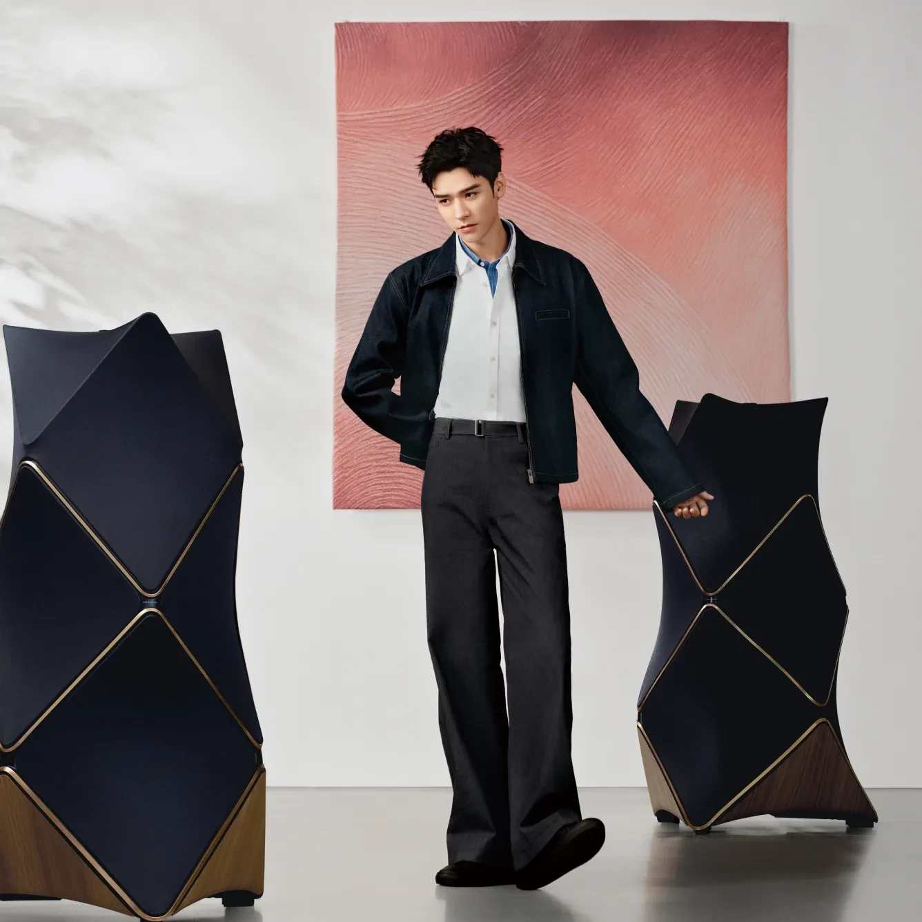 Person in dark jacket and white shirt posing against pink textured backdrop with geometric navy blue Beolab 90 floor speakers.