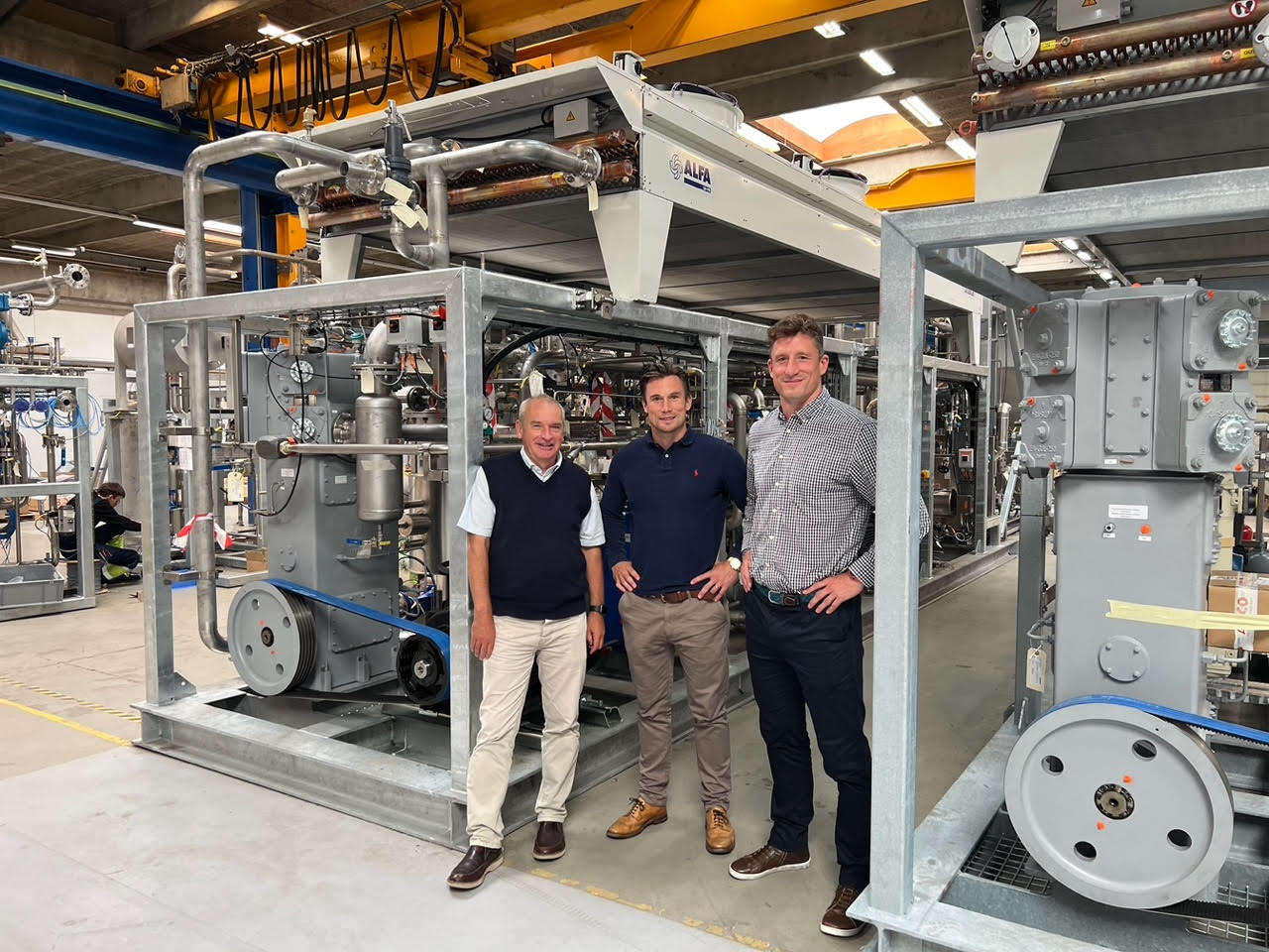 Three men standing in front of large industrial machinery in a factory or workshop setting.