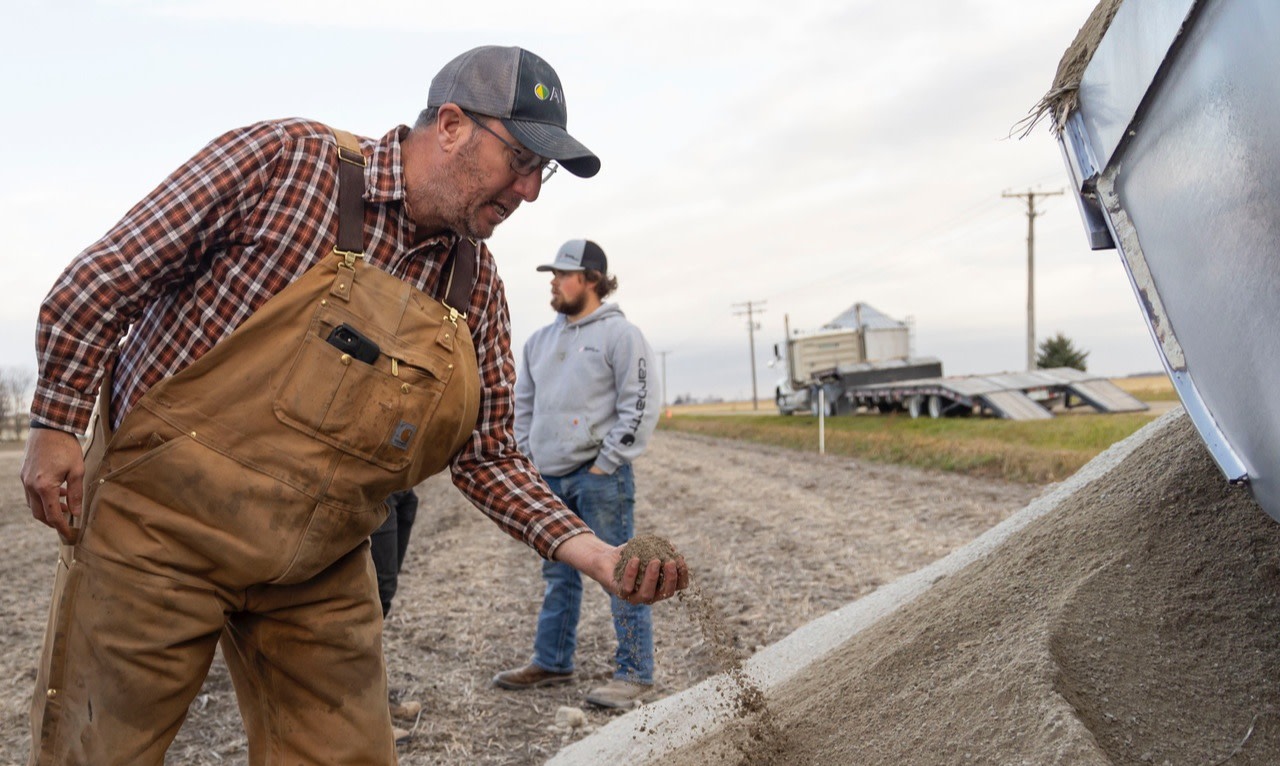 Two farmers in rural setting, one examining grain pouring from truck, other watching. Grain silo visible in background.