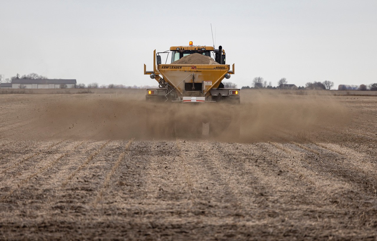 Yellow agricultural spreader driving across a bare field, kicking up dust as it distributes material on the soil.