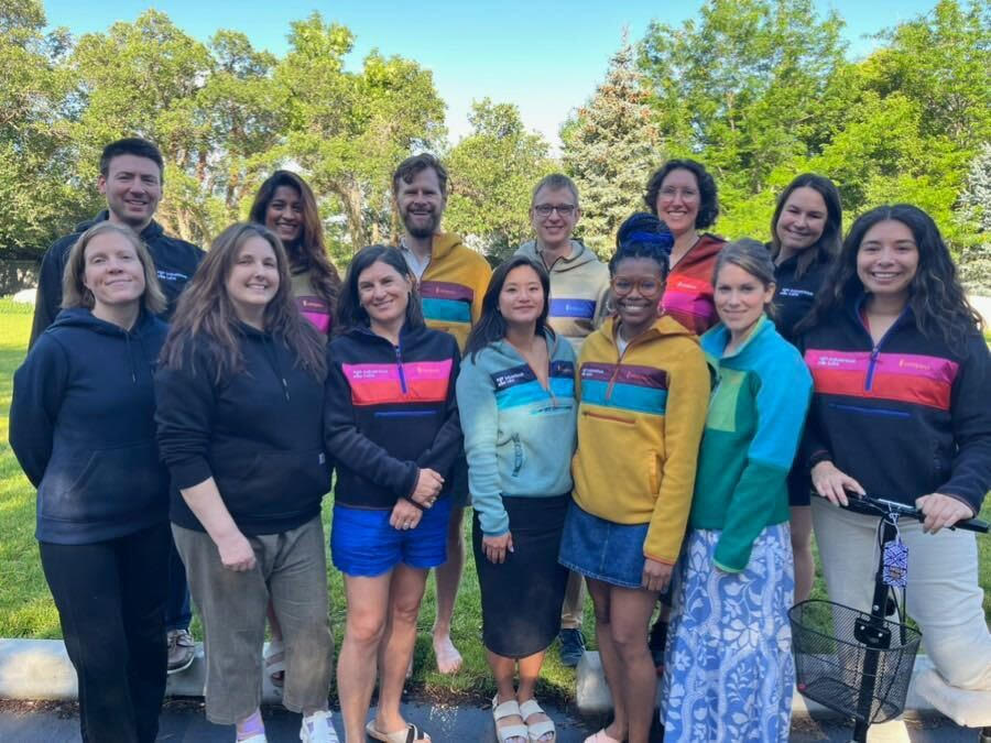 A diverse group of people standing together outdoors in a park, wearing colorful casual clothing and smiling at the camera.