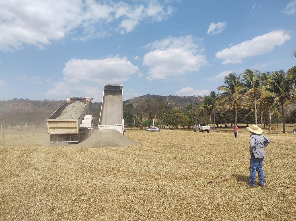 Dump truck unloading gravel on dry field, worker observing. Palm trees and hills in background under blue sky with clouds.