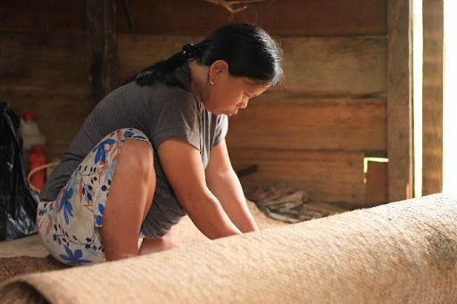 Woman working on a large textile or rug in a rustic wooden interior, focused on her craft.