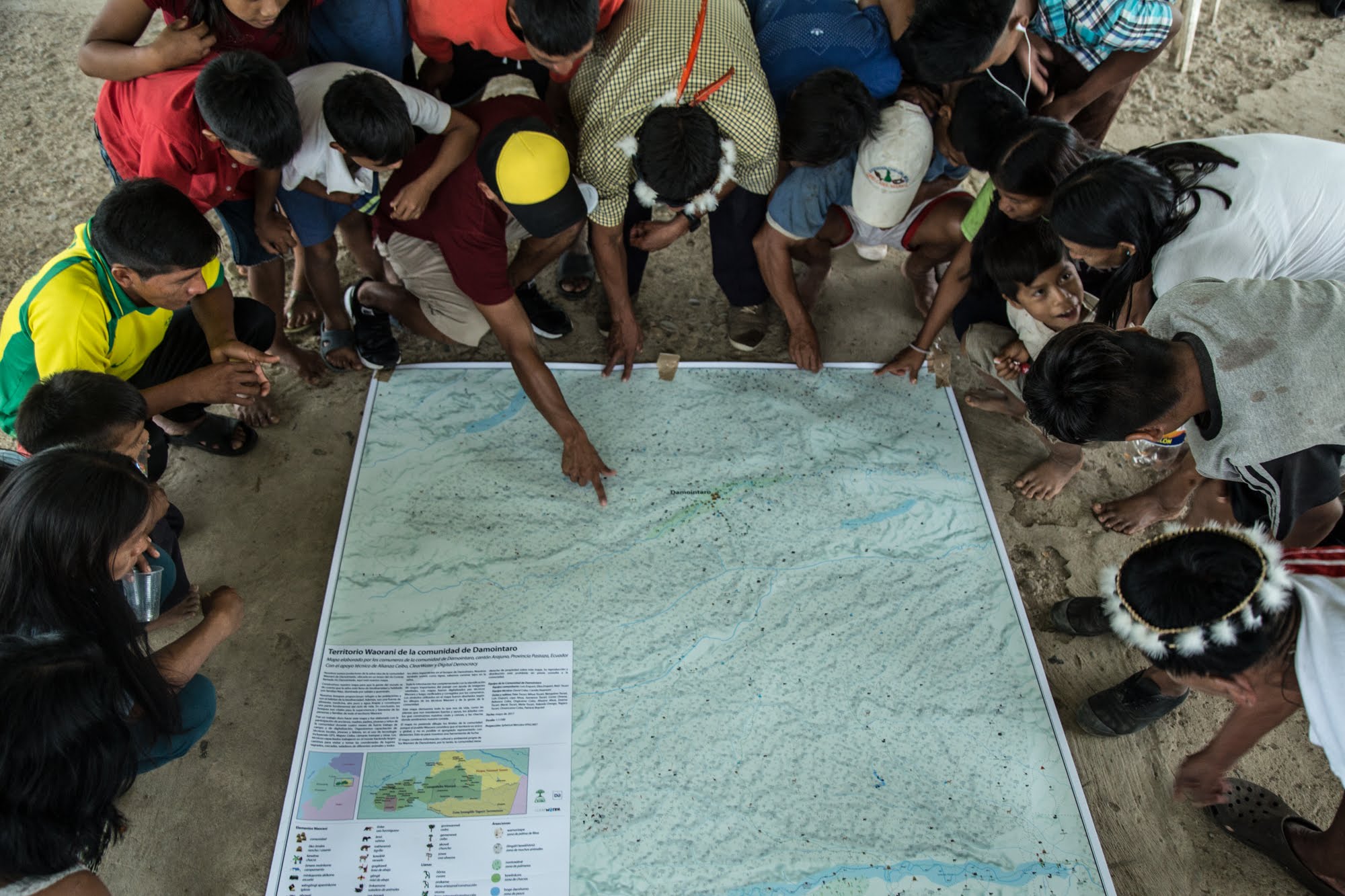 Group of children gathered around a large map on the ground, with one child pointing at a location. Colorful clothing visible.