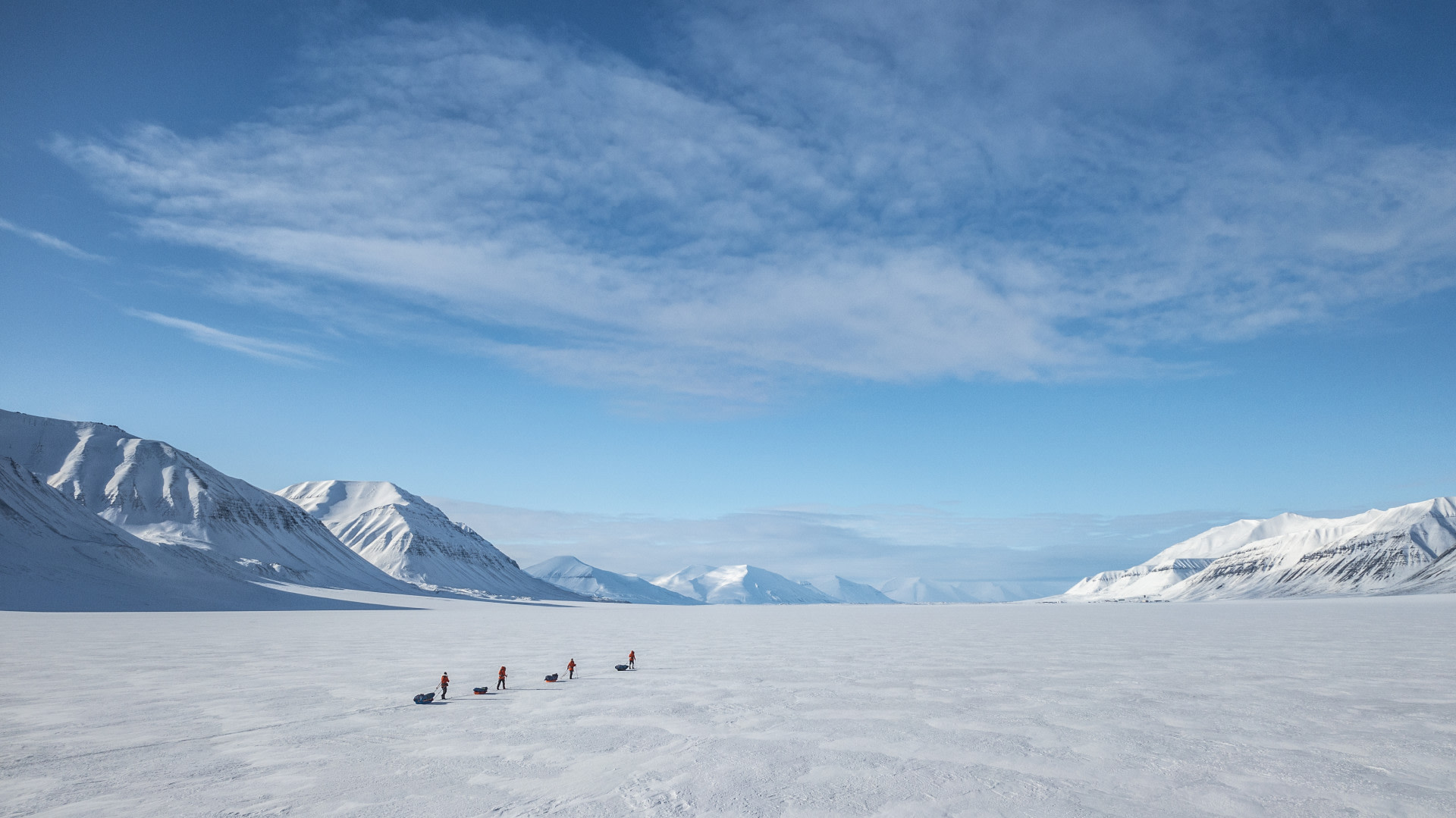 Dog sled team traversing a vast snowy plain, surrounded by majestic snow-capped mountains under a bright blue sky with wispy clouds.