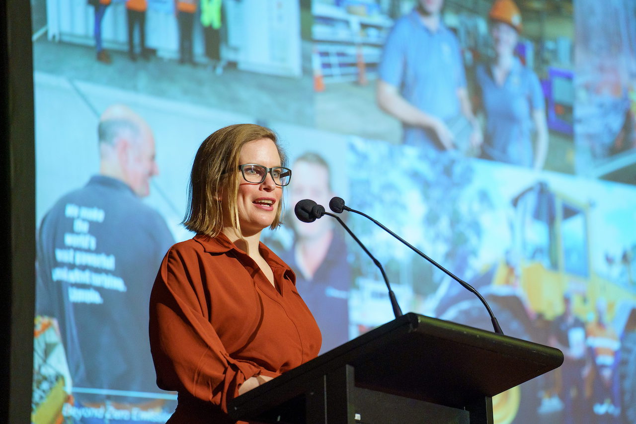 Woman in glasses and rust-colored shirt speaking at podium with large projection screen behind showing crowd scene