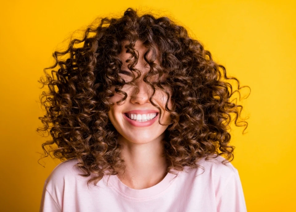 woman with 3a curly hair smiling with her eyes closed as she stands in front of a yellow background 