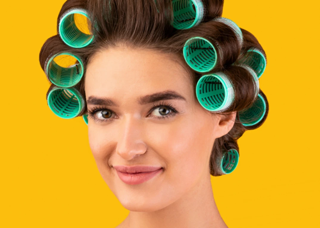 A woman with green heated rollers in her brown hair looks forward in front of a yellow background.