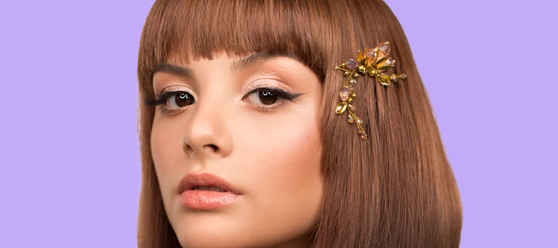 A woman with short brown bangs and a hair clip looks forward in front of a purple background.