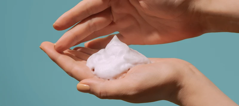 a person with fair skin holds mousse in the palm of their left hand in front of a gray blue background.