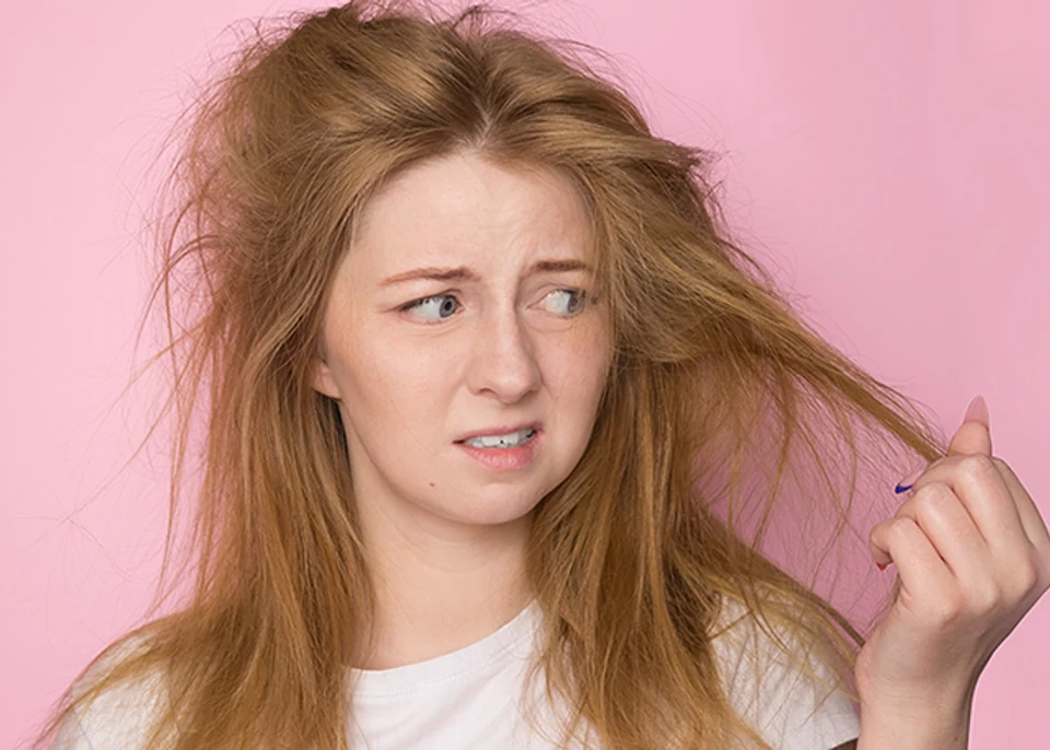 lifeless sun damaged hair. A woman on a pink background holds her disheveled, tangled hair and looks at it with a discontented expression.