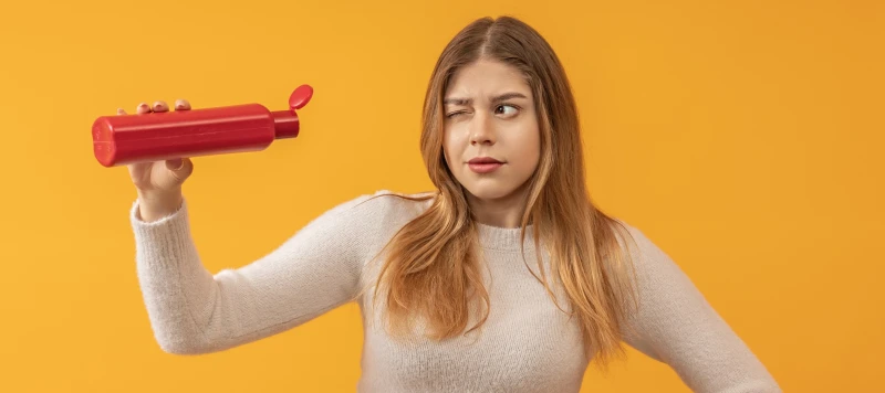 a young woman with long hair looks with one eye into an open bottle, find out if the shampoo is empty or not, color background