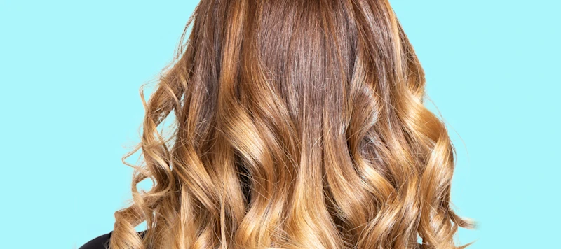  A person with a brown and blonde balayage hairstyle faces away in front of a light blue background.