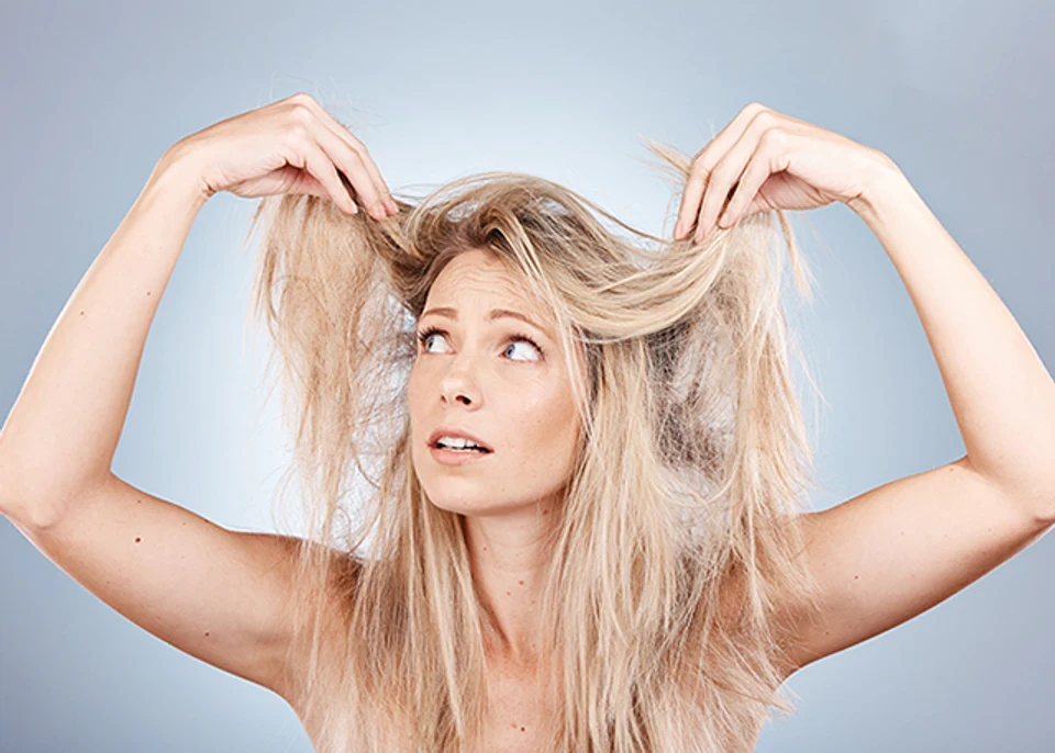 Blonde woman holding up her hair and frustrated with the frizz, against a grey background