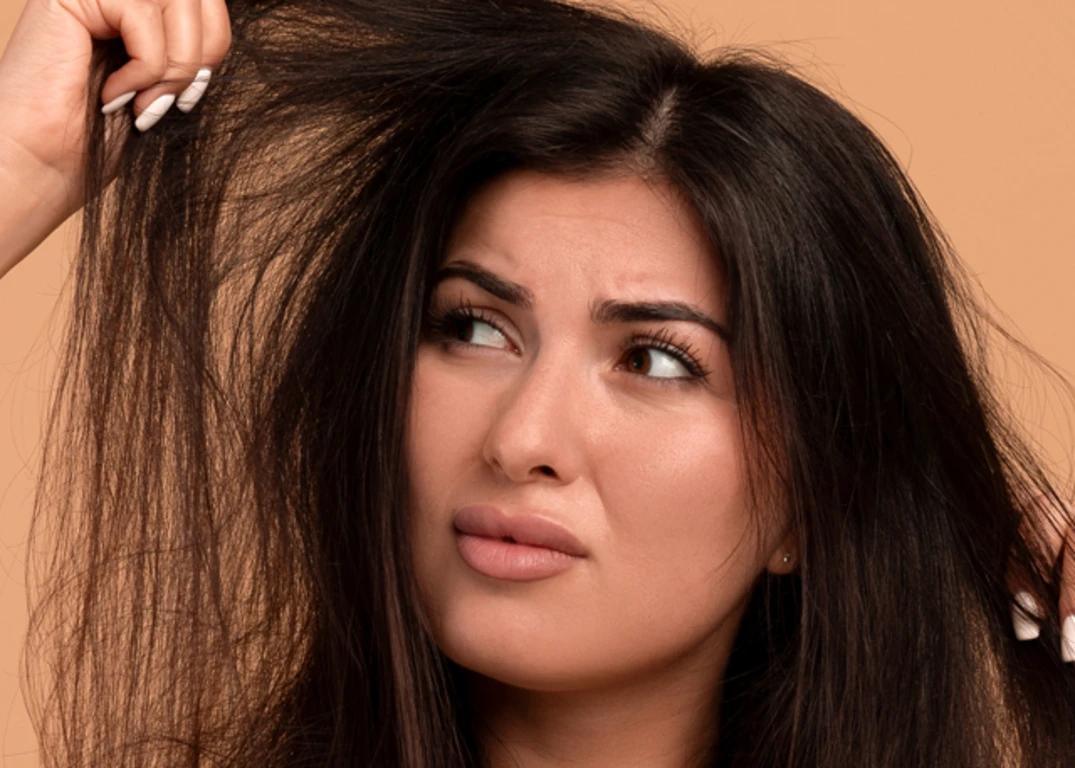 Woman with long brown frizzy hair holds it up and looks at it intently in front of a light brown background.