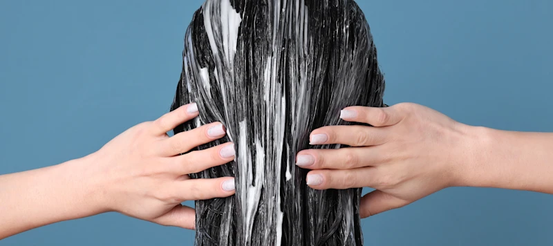 Fair skinned woman’s hands touch her black hair coated with hair conditioner in front of a blue background