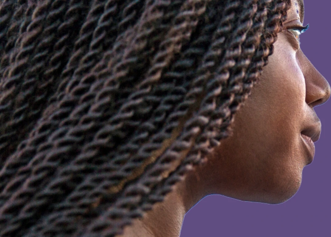 A dark-skinned woman with hair in a senegalese twist looks to the right in front of a purple background. 
