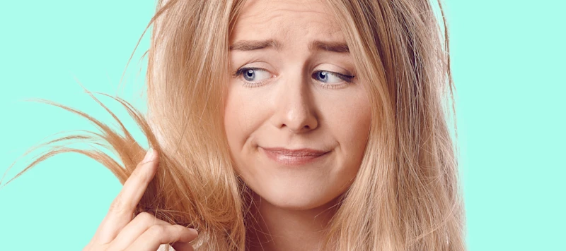 a fair skinned woman with damaged bleached hair holds it up with her fingers at looks at it with concern.