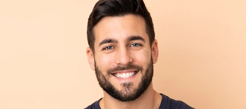 a man with fair skin, brown hair and a beard smiles looking forward in front of a light orange background.