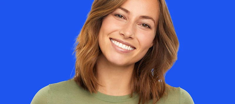 A woman with fair skin and medium length light brown smiles facing forward in front of a blue background.