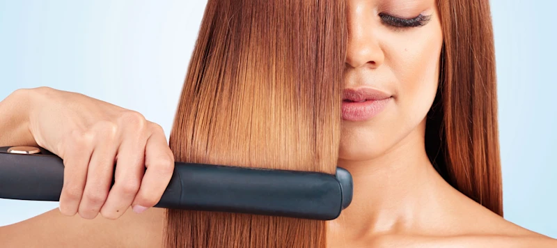 Woman with tan skin and yellow brown hair uses flat iron to straighten her hair in front of a grey background
