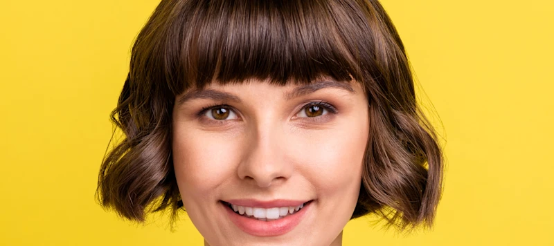 a woman with fair skin and a short brown bob hairstyle with bangs looks forward and smiles in front of a yellow backgound