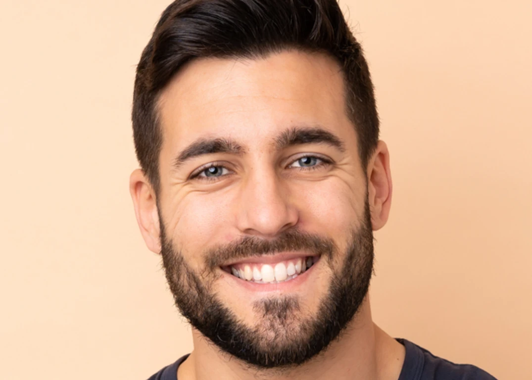 a man with fair skin, brown hair and a beard smiles looking forward in front of a light orange background.