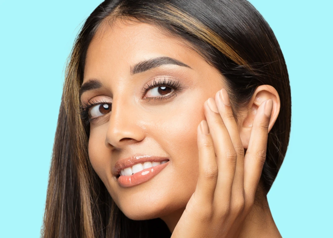 A woman with tan skin and highlights in her dark brown hair touches her face and looks forward.