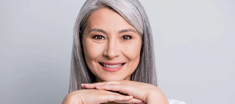 A woman with long gray hair smiles facing forward with her hands below her chin in front of a white background.