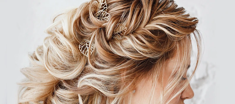 A woman with fair skin, wavy blonde hair in a prom hairstyle with leaf shaped clips looks down and right in front of a white background.