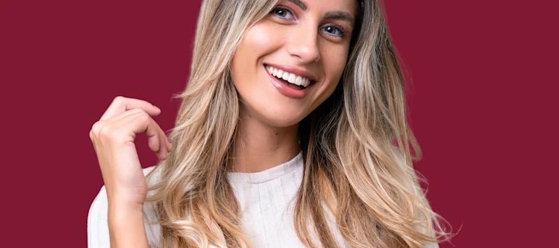 A woman with fair skin and demi permanent blonde hair smiles facing forward in front of a maroon background.