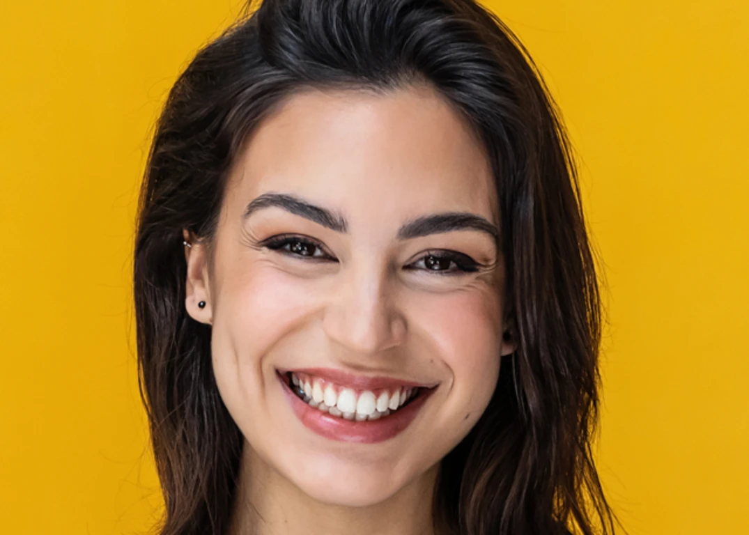 Woman with olive complexion dark eyes and dark hair smiles forward in front of a yellow background.