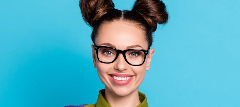 A woman withy fair skin, glasses and two dark brown space buns smiles forward in front of a light blue background.