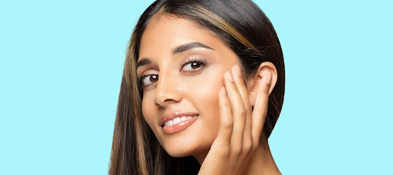 A woman with tan skin and highlights in her dark brown hair touches her face and looks forward.