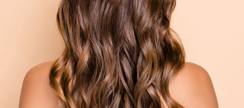 A woman with brown heat protected hair faces backwards in front of a light brown background.