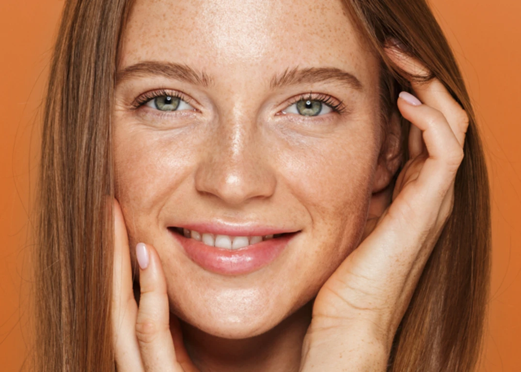 Woman with round face and thin light brown straight hair smiles looking forward in front of an orange background.