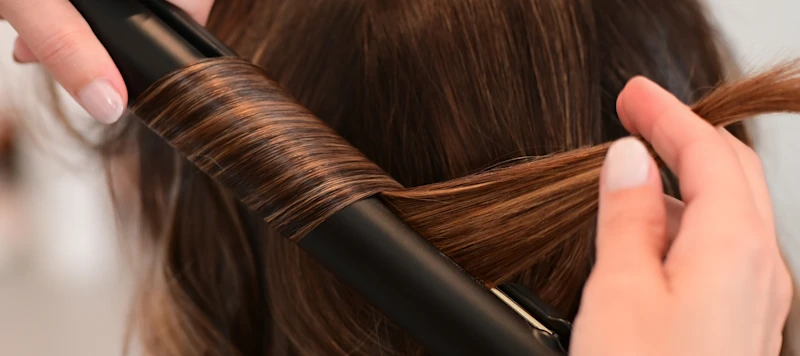 A stylist uses a black flat iron to curl a woman’s brown hair.