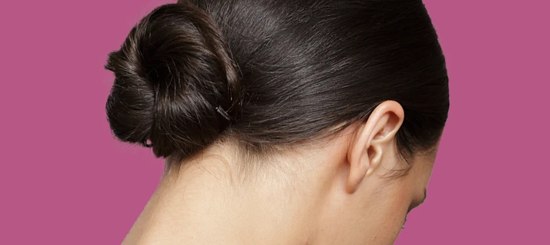 A woman with fair skin and dark brown hair in a bun looks down in front of a magenta background.