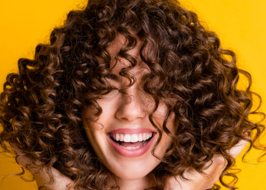 A woman with curly brown hair holds it up with flat hands in front of a yellow background.