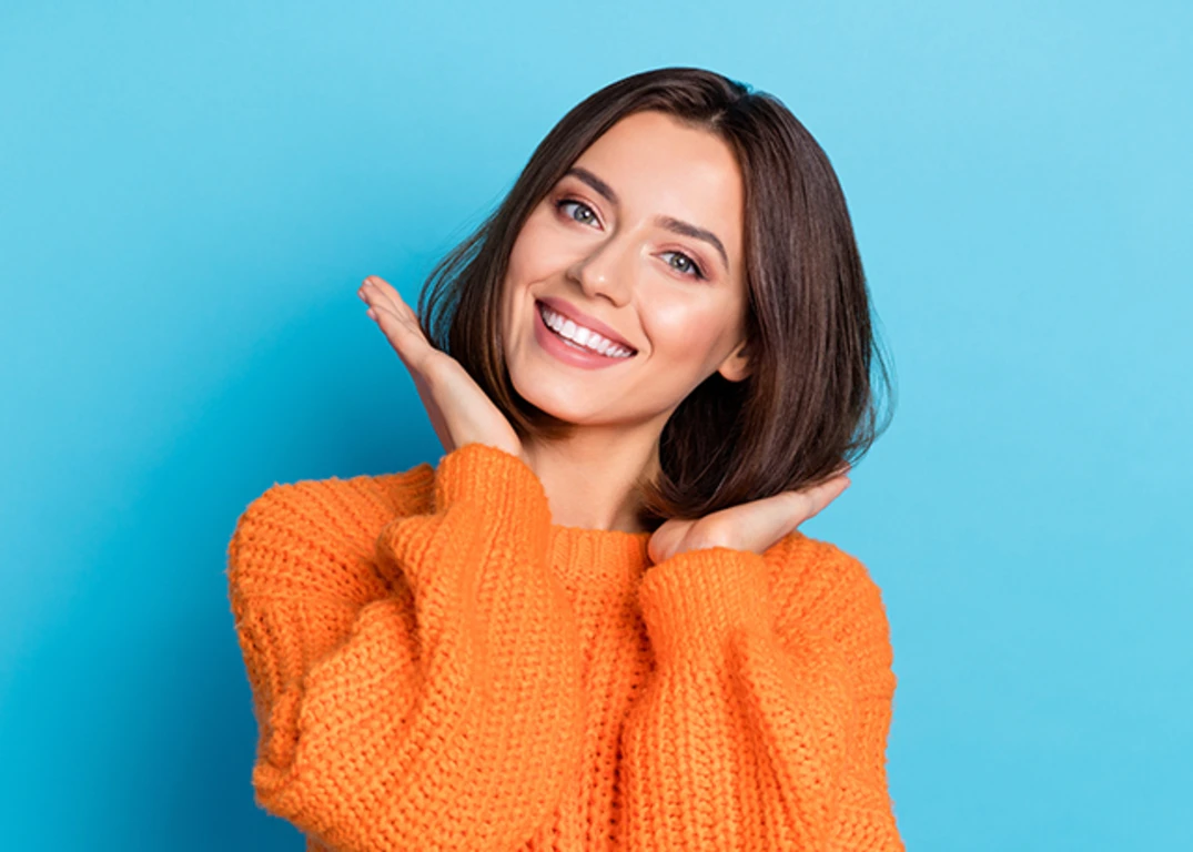woman wearing orange sweater smiling touching the ends of her brown well-groomed hair