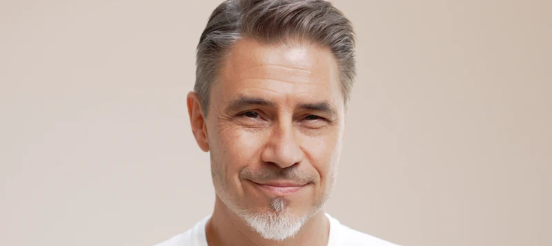 Senior man with fair skin and short gray hair smiles in front of a light brown background.