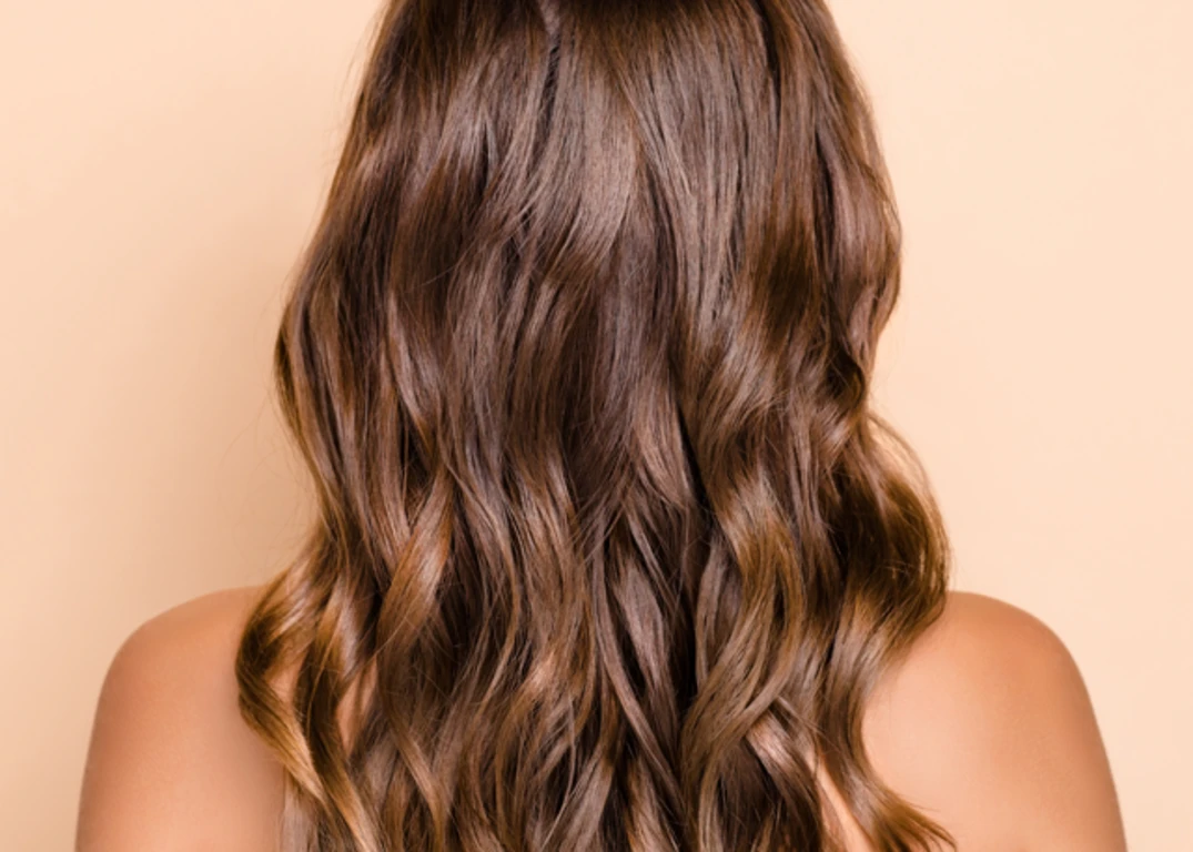 A woman with brown heat protected hair faces backwards in front of a light brown background.